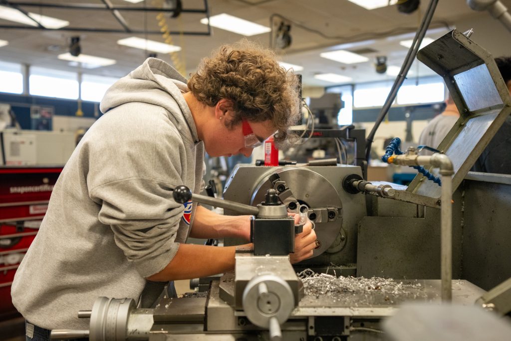 Un estudiante trabaja con un torno en un taller, concentrado en lo que hace y con gafas de seguridad puestas. Las virutas se esparcen por todas partes.