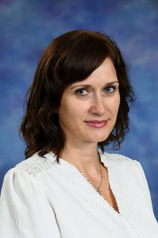 A woman with shoulder-length dark hair, wearing a white blouse, smiles against a blue background.