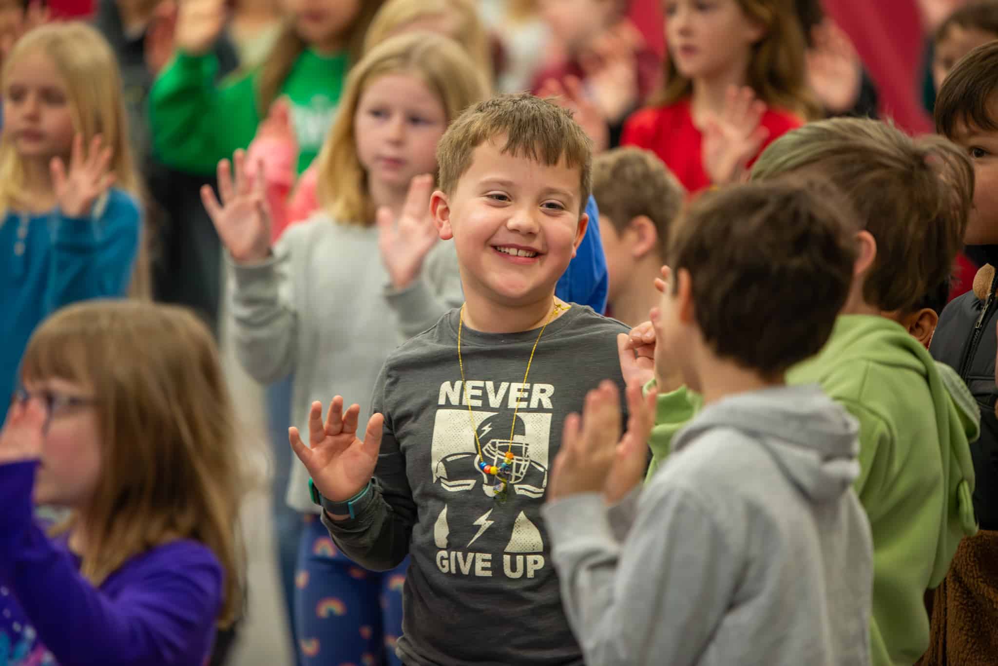 3rd grade boy standing in a crowd with both of his elbows bent so his hands are in front of his shoulders while he practices movement for a performance. He is smiling. Kids around him are in the same position.