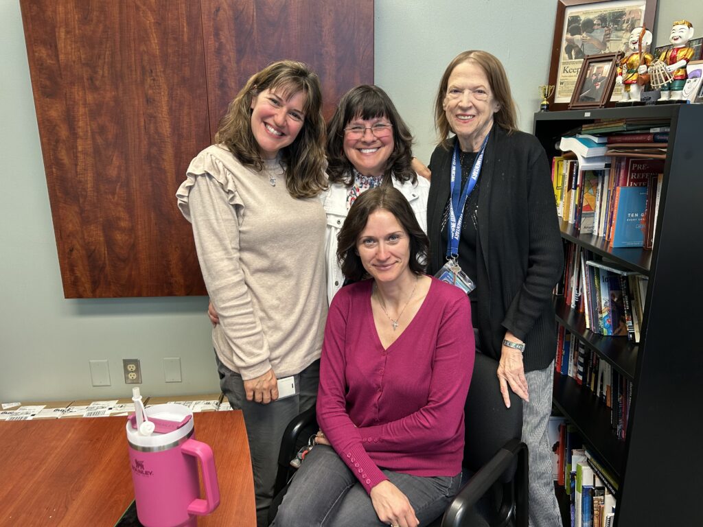 Four women pose together in an office setting with shelves of books behind them, smiling and looking cheerful.