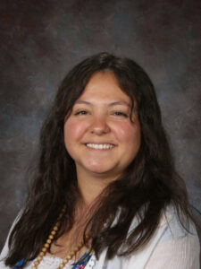 Smiling woman with long dark hair, wearing a necklace and a white top, poses against a textured dark background.