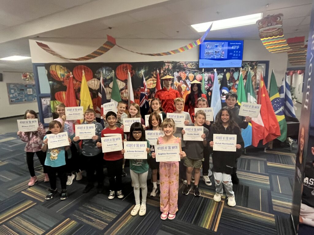 A group of students in a school celebrating "Student of the Month," holding certificates with flags in the background.