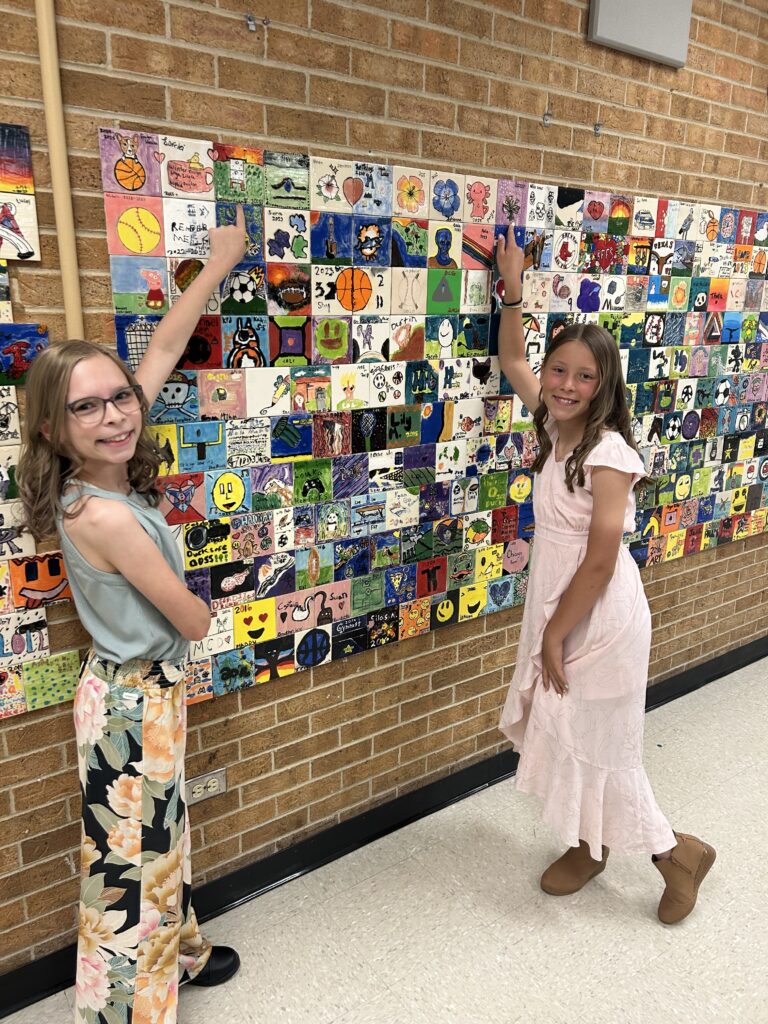 Two girls stand in front of a colorful art wall, pointing at various painted square tiles depicting different designs.