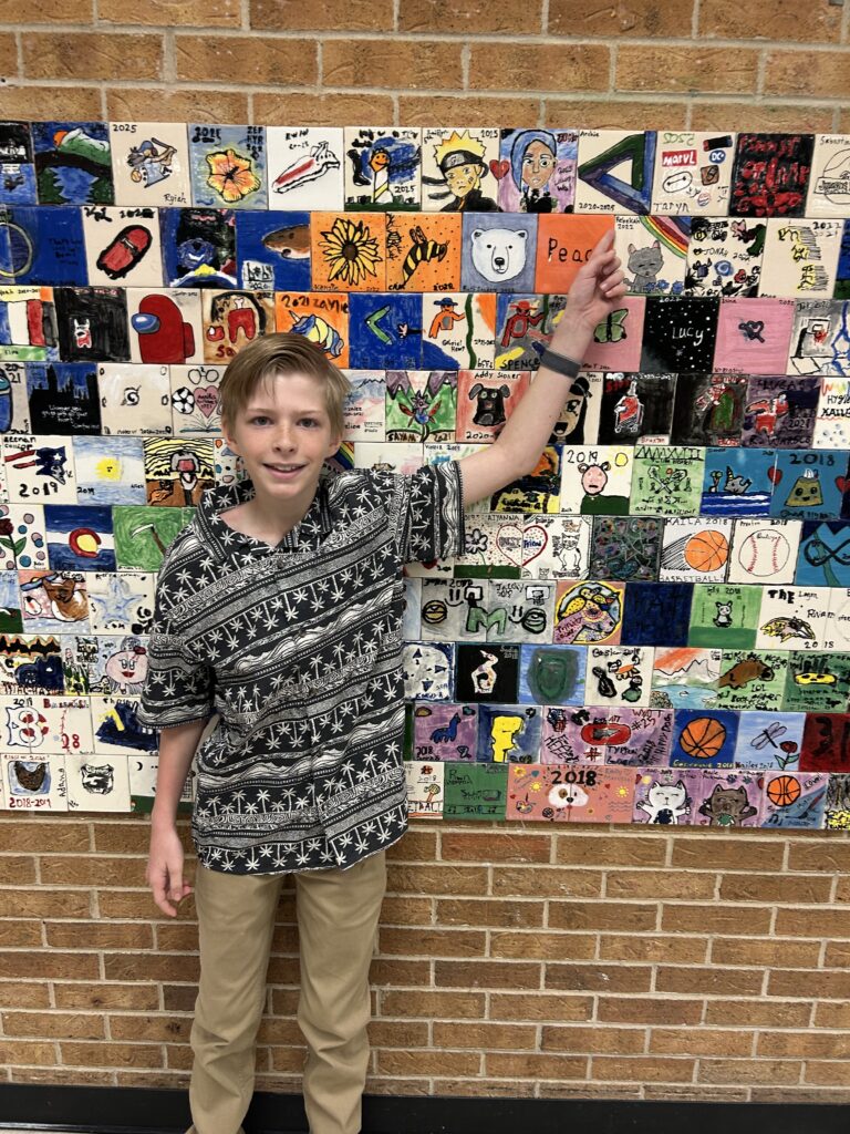 A boy stands in front of a colorful mural made of various painted tiles, pointing to one titled "Peace."