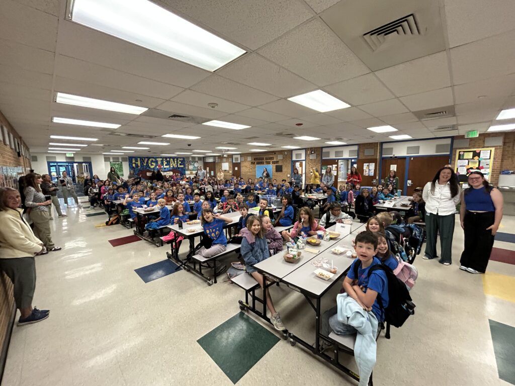 A school cafeteria filled with students in blue shirts, sitting at tables, smiling, and enjoying lunch together.