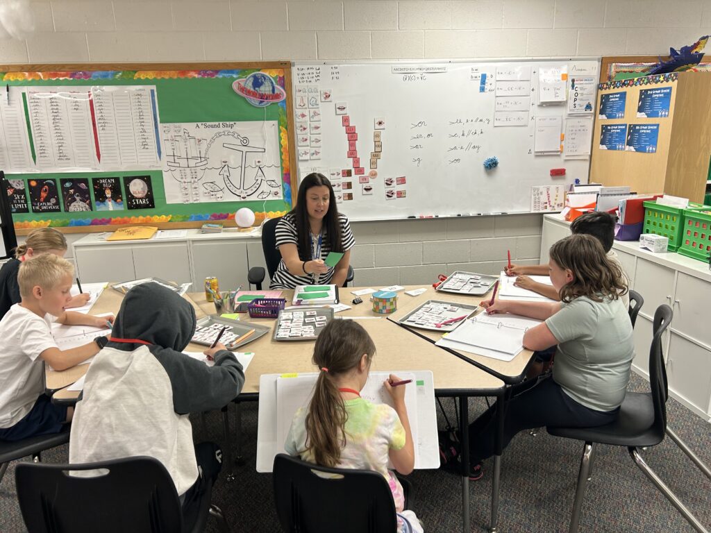A teacher instructs a small group of students at a classroom table, as they work on assignments and take notes.