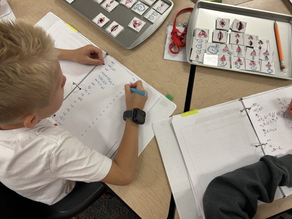 A child is writing in a notebook, surrounded by learning materials, including a tray of educational cards.