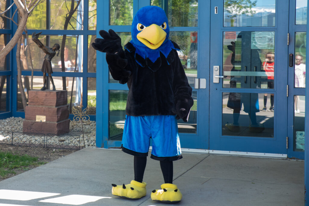 A blue bird mascot in black and blue attire waves near a building entrance, with a statue visible in the background.