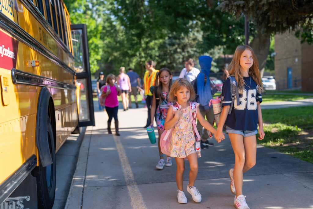 Children walking from a school bus, with two girls holding hands in the foreground and others in the background.