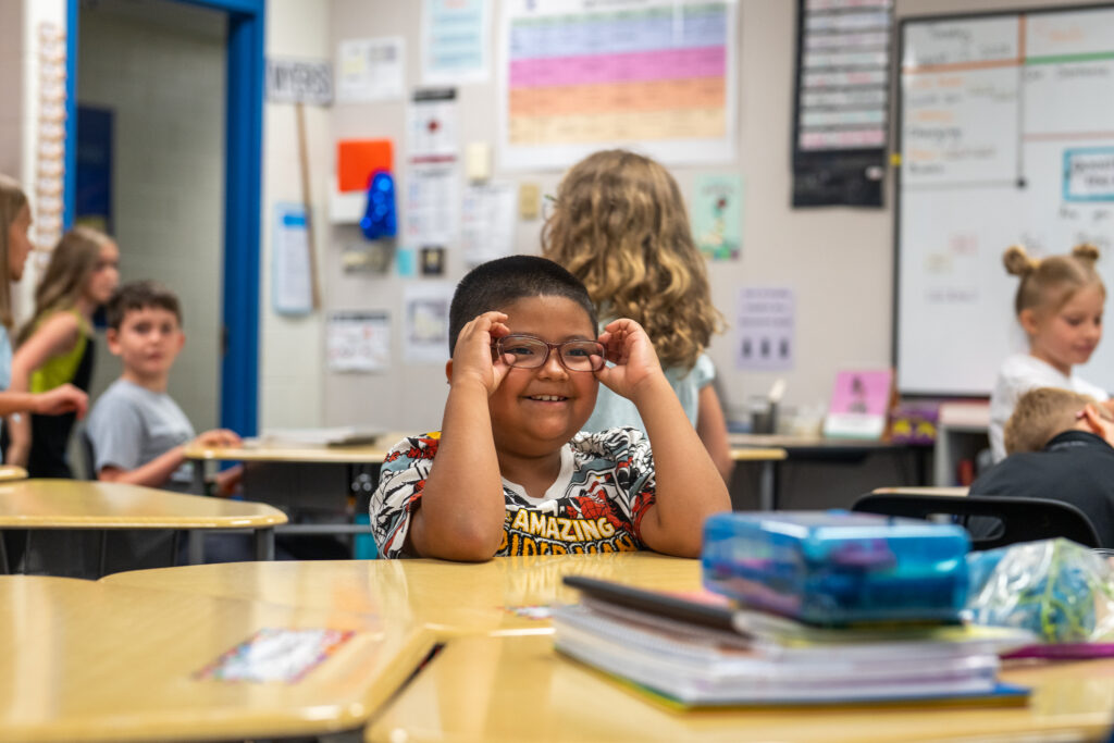 A smiling child with glasses sits at a classroom desk, while other students are engaged in activities in the background.