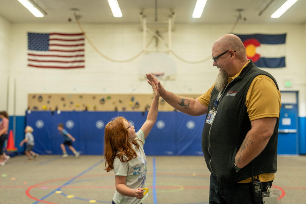 A young girl high-fives a smiling man in a gym, with an American flag and Colorado flag in the background.