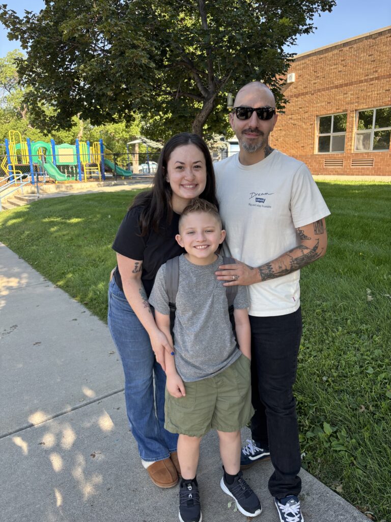 A family of three poses for a photo outside near a playground, smiling together with green grass in the background.