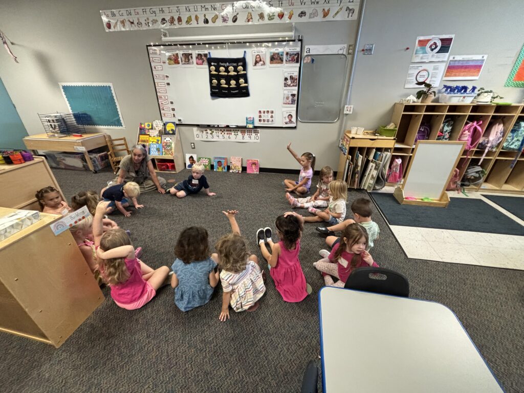 A group of young children sits on the floor in a classroom, engaged with an adult instructor.