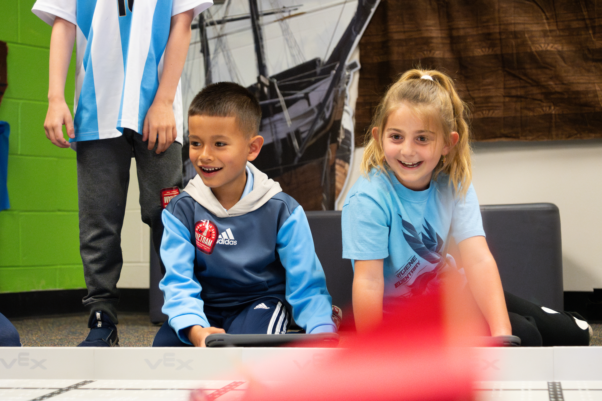 Two children, one boy and one girl, smile while engaging with a robotics activity in a classroom setting.