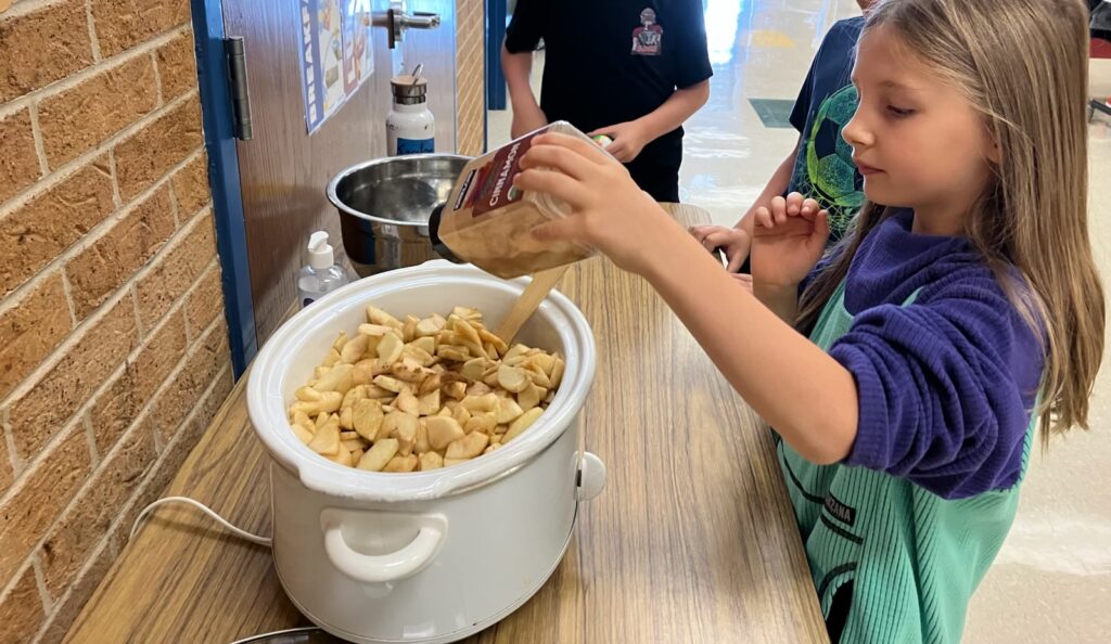 A child pours cinnamon over sliced apples in a slow cooker while others assist nearby. A clean, bright kitchen setting.