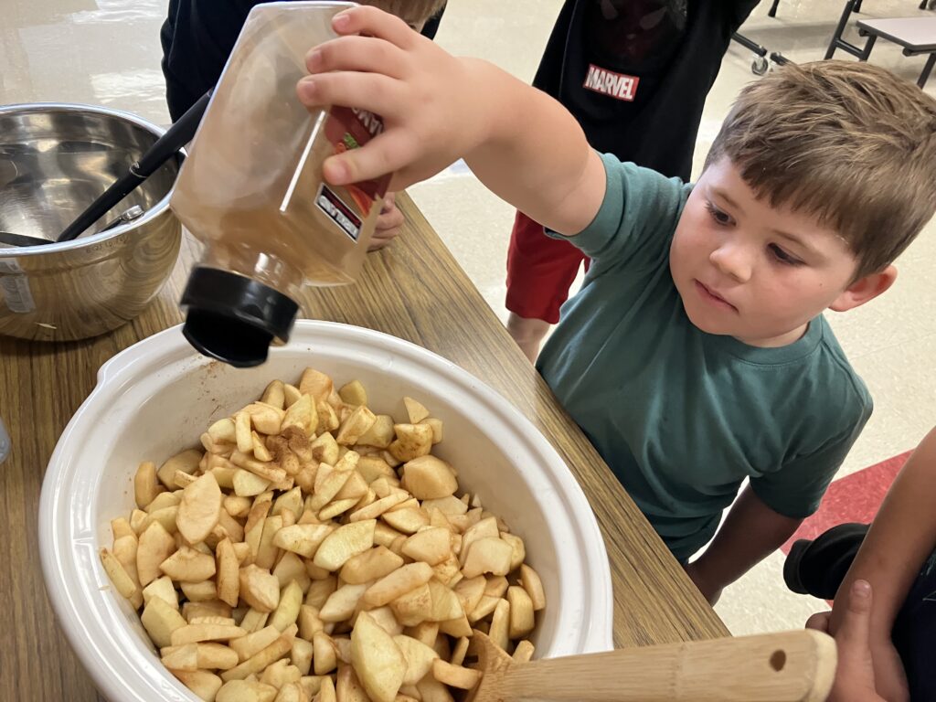A child pours cinnamon over a bowl of sliced apples while focused on the task, with a mixing bowl in the background.