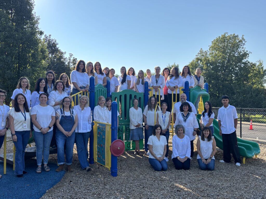 A group of educators in white shirts pose together on a playground, surrounded by trees and clear blue skies.