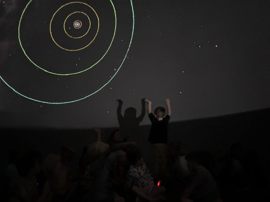 A child raises their arms in excitement in front of a starry projection with colorful circular orbits.