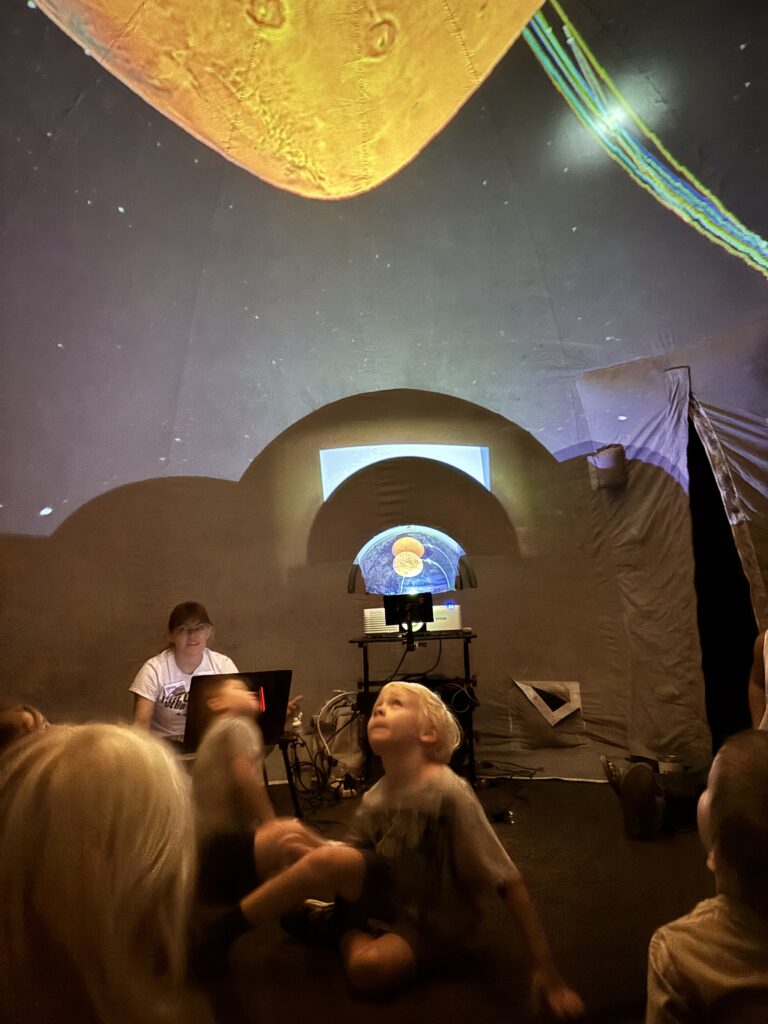 Children sit on the floor, looking up at space projections while an adult presents from a laptop.