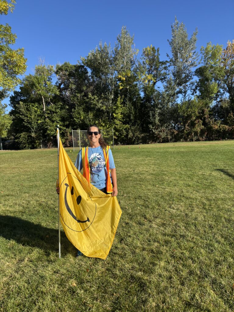 A person in a yellow safety vest holds a large yellow flag with a smiley face, standing on a grassy field.