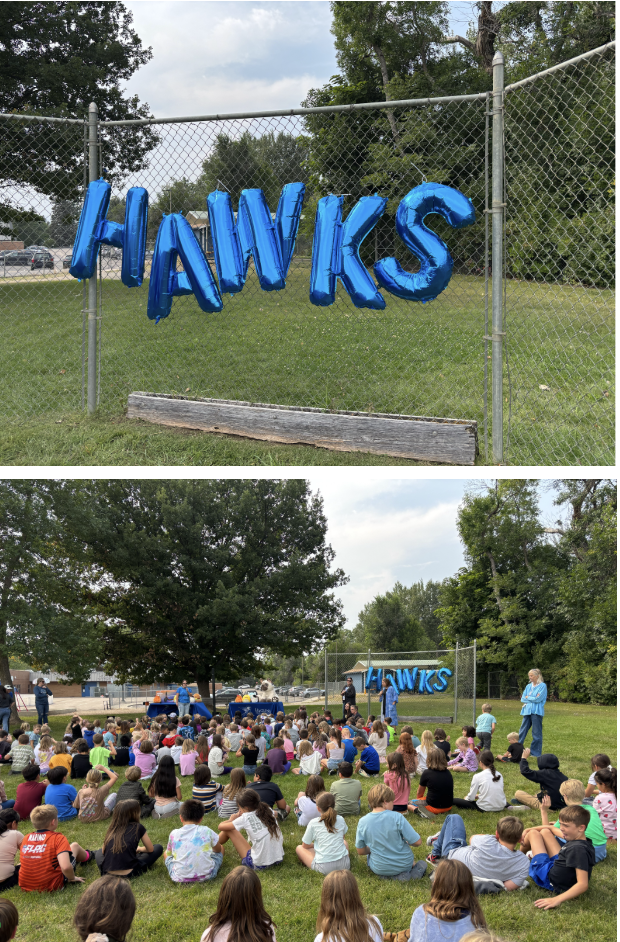 Blue balloons spelling "HAWKS" are displayed on a fence with students sitting on the grass for an event.
