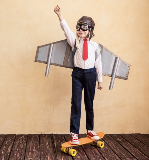 A child in an aviator outfit stands on a skateboard, wearing goggles and a cardboard jetpack, striking a heroic pose.