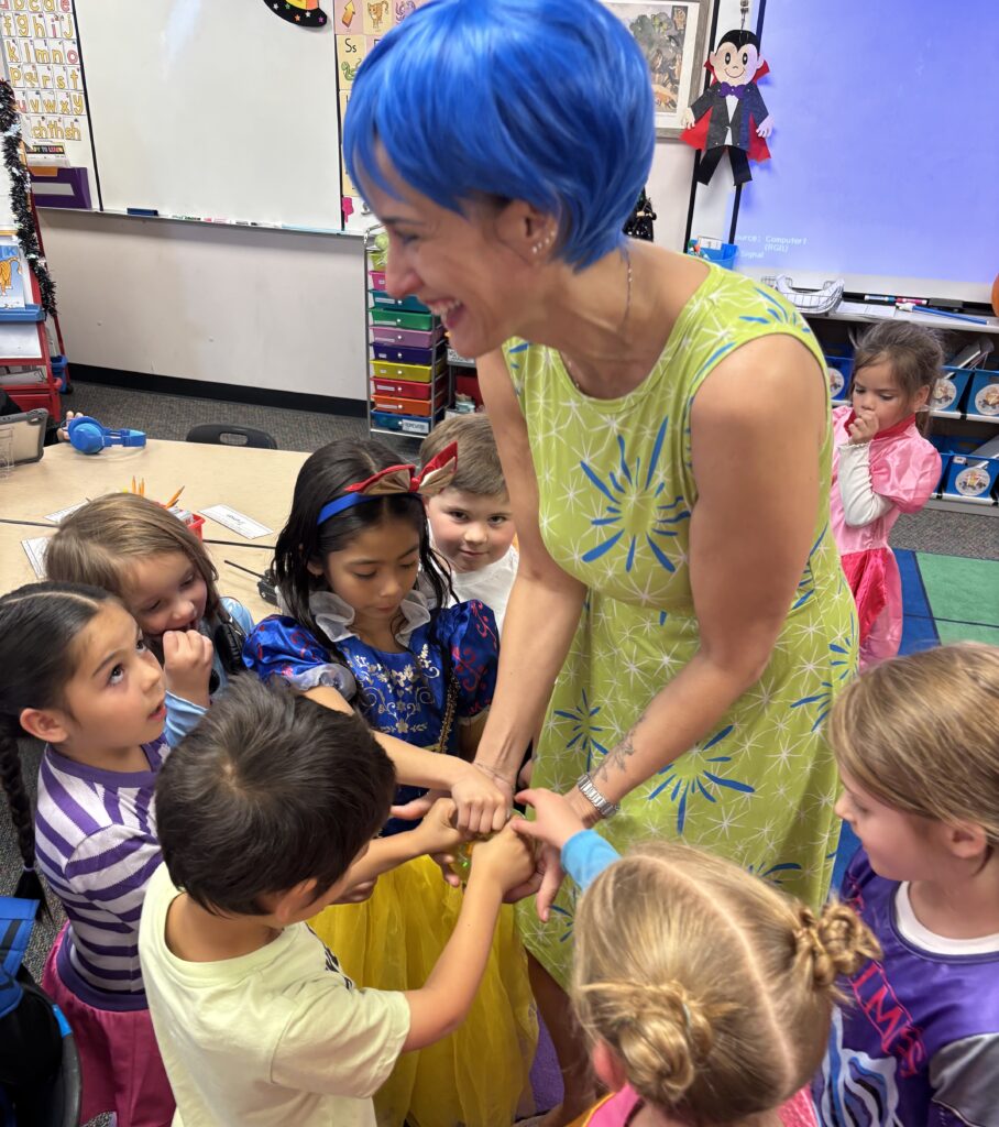 A teacher with blue hair interacts joyfully with a group of children in colorful costumes in a classroom setting.