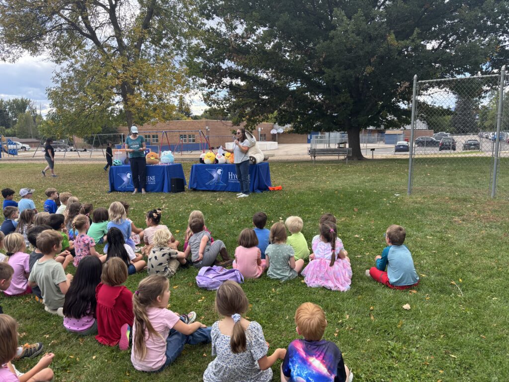 Children sit on the grass watching a presentation with colorful decorations and speakers at an outdoor school event.