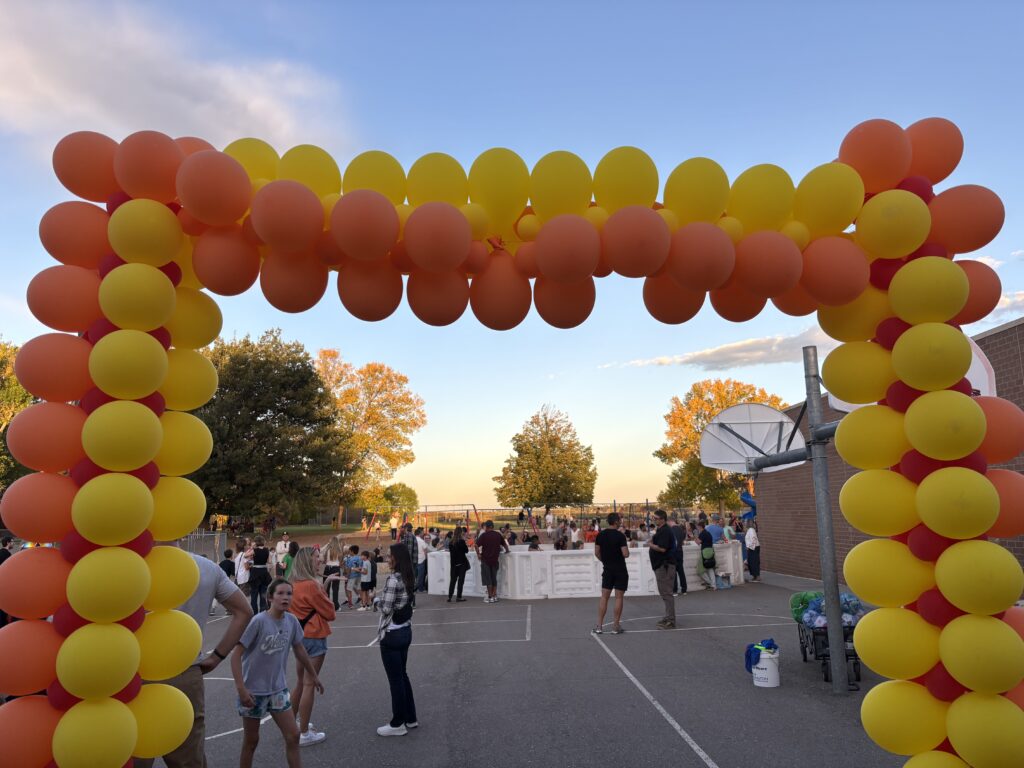 A colorful balloon arch frames a busy outdoor gathering with people, trees, and playground equipment in the background.