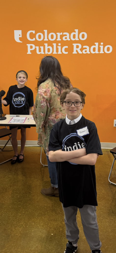 Two children in black "indie 102.3" shirts at Colorado Public Radio, one smiling at the camera, the other at a table.