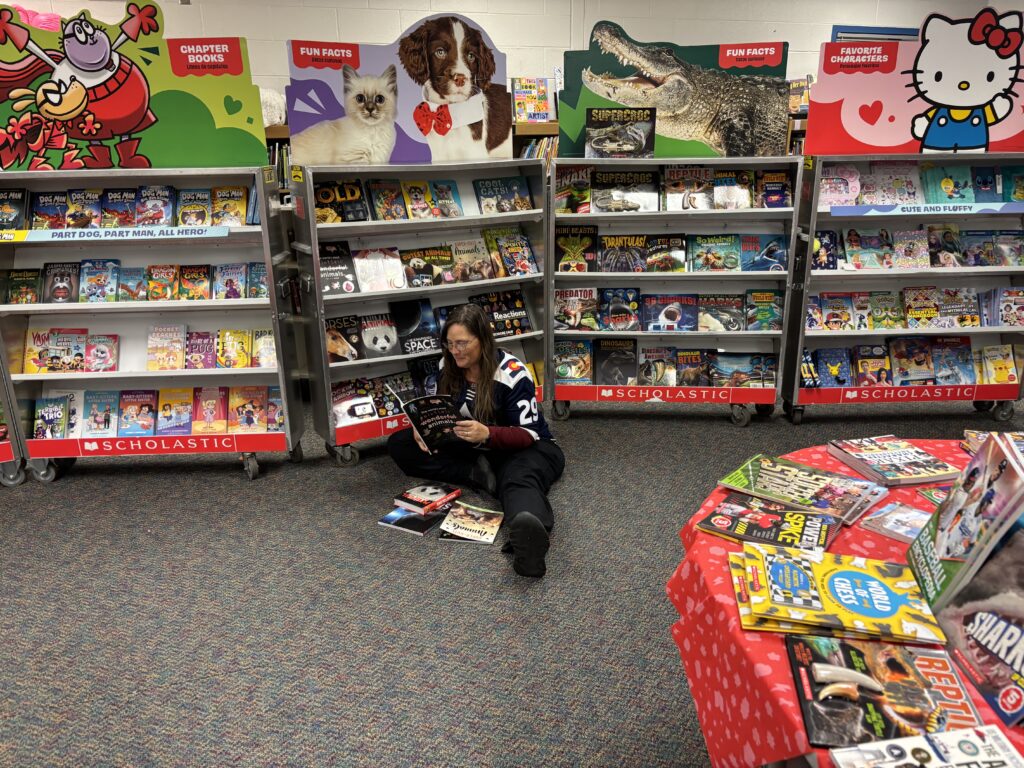 A person sits on the floor of a colorful book fair, reading a book surrounded by shelves of children's books.