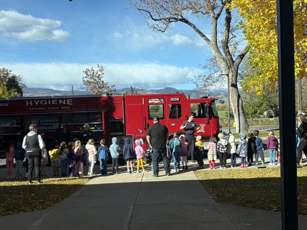 Children gather around a red fire truck, listening to adults while trees with autumn leaves are in the background.