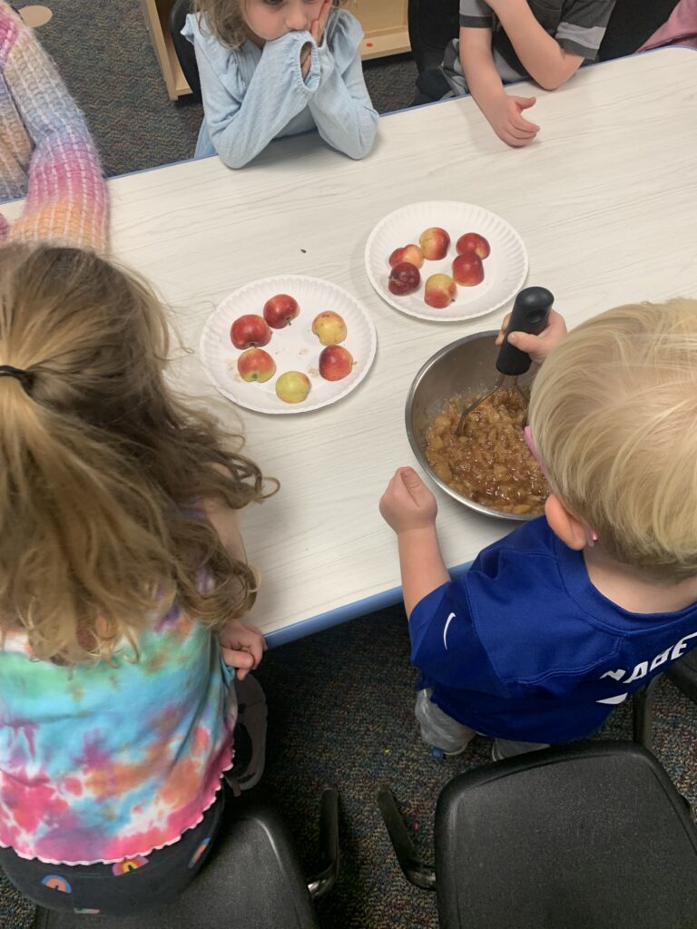 Children are gathered around a table with apples and a bowl of apple mixture, engaging in a cooking activity.
