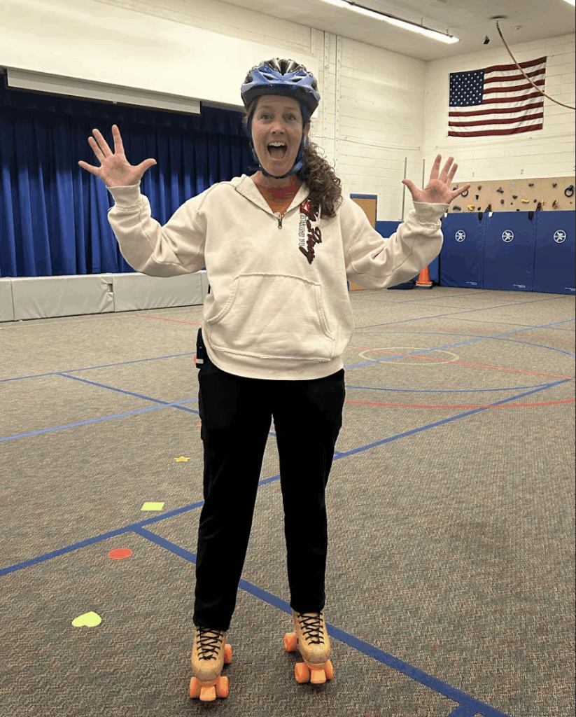 A woman in a helmet and roller skates smiles widely, standing in a gym with colorful floor markings.