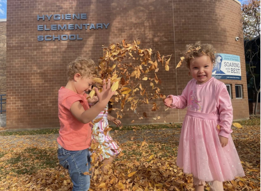 Children play in a pile of autumn leaves outside Hygiene Elementary School, joyfully tossing leaves in the air.