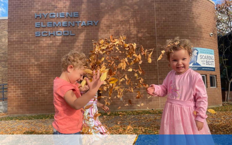 Two young children play with autumn leaves in front of Hygiene Elementary School, smiling and enjoying the moment.
