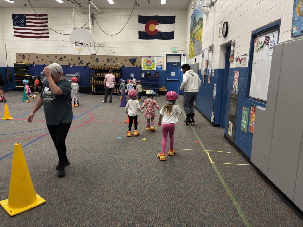 Children roller skating indoors, with safety cones and adults supervising in a gym decorated with flags and colorful artwork.