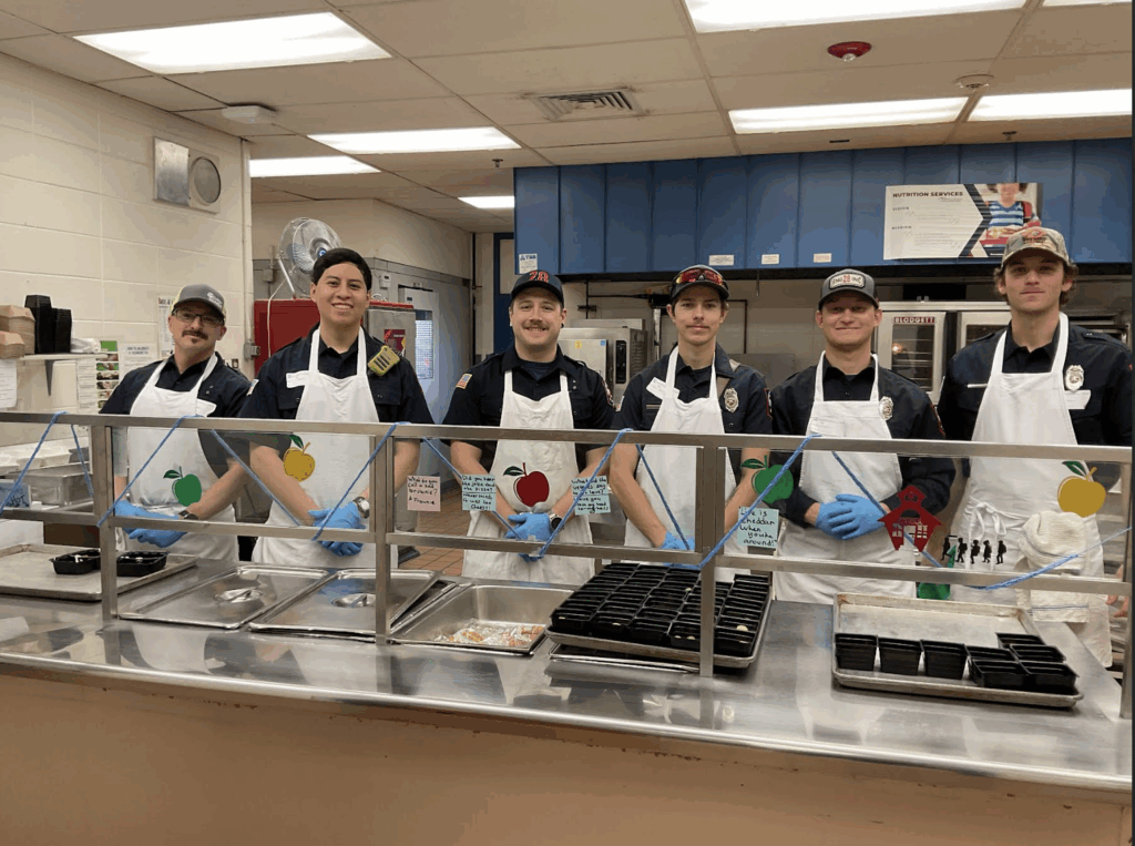 Six uniformed individuals stand behind a food service counter in a kitchen, wearing aprons and gloves.