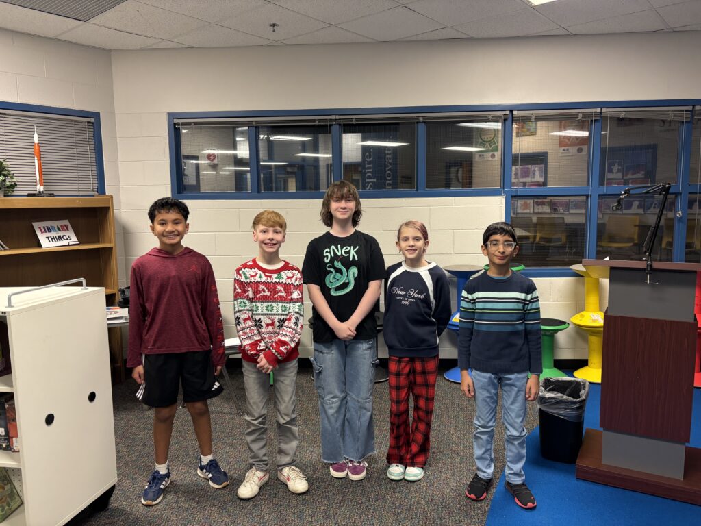 Five children stand together in a library, smiling. A bookshelf and a podium are visible in the background.