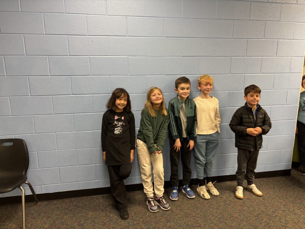 Five children stand in a line against a light blue wall, smiling and wearing casual clothing. A chair is nearby.