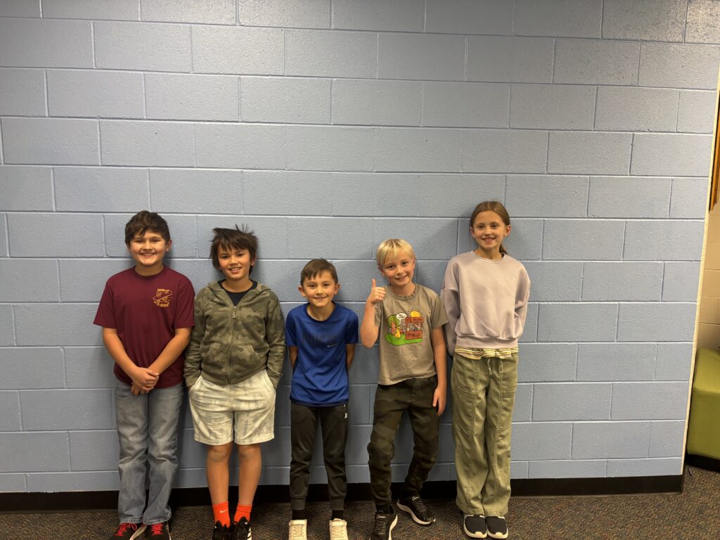 Five children stand in a line against a light blue wall, smiling and dressed in casual clothing.
