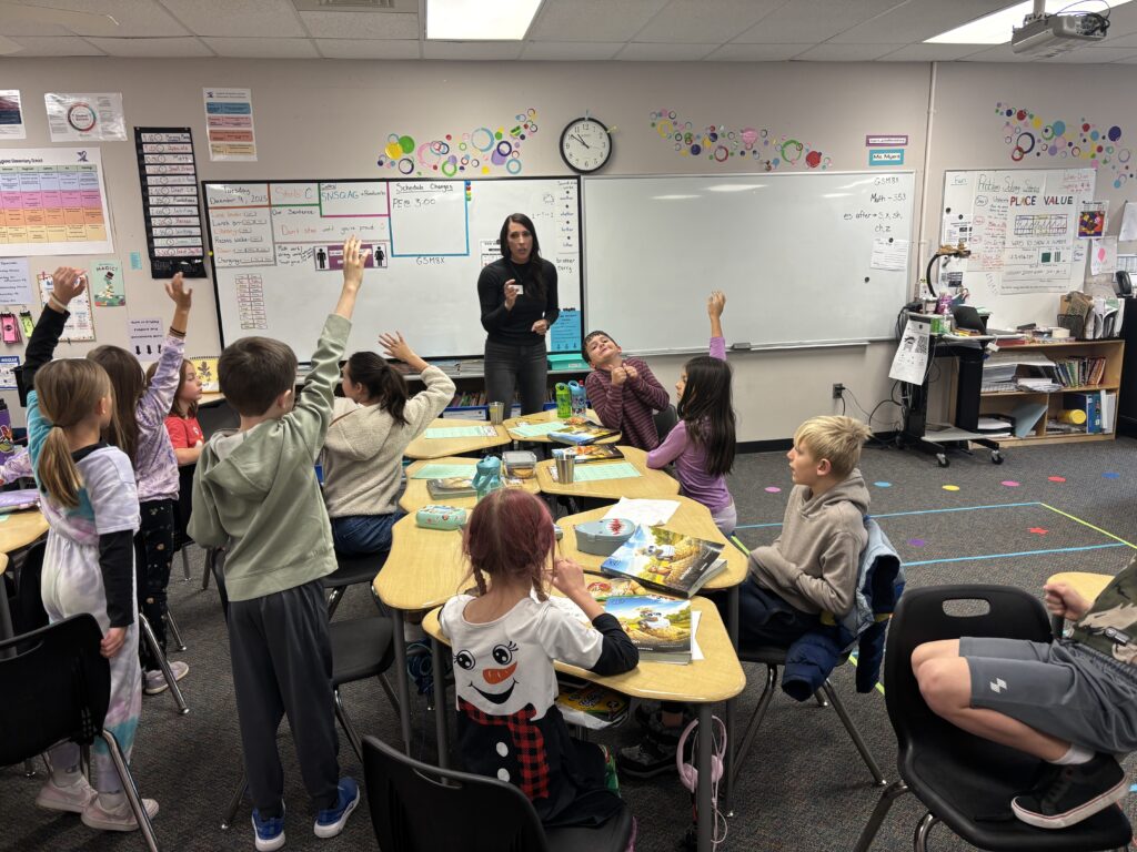 A teacher instructs a classroom of students, most with raised hands, engaged in a lesson with various materials on desks.