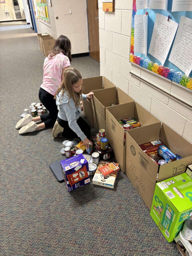 Two children are organizing food items into cardboard boxes in a school hallway. Papers are displayed on the wall.