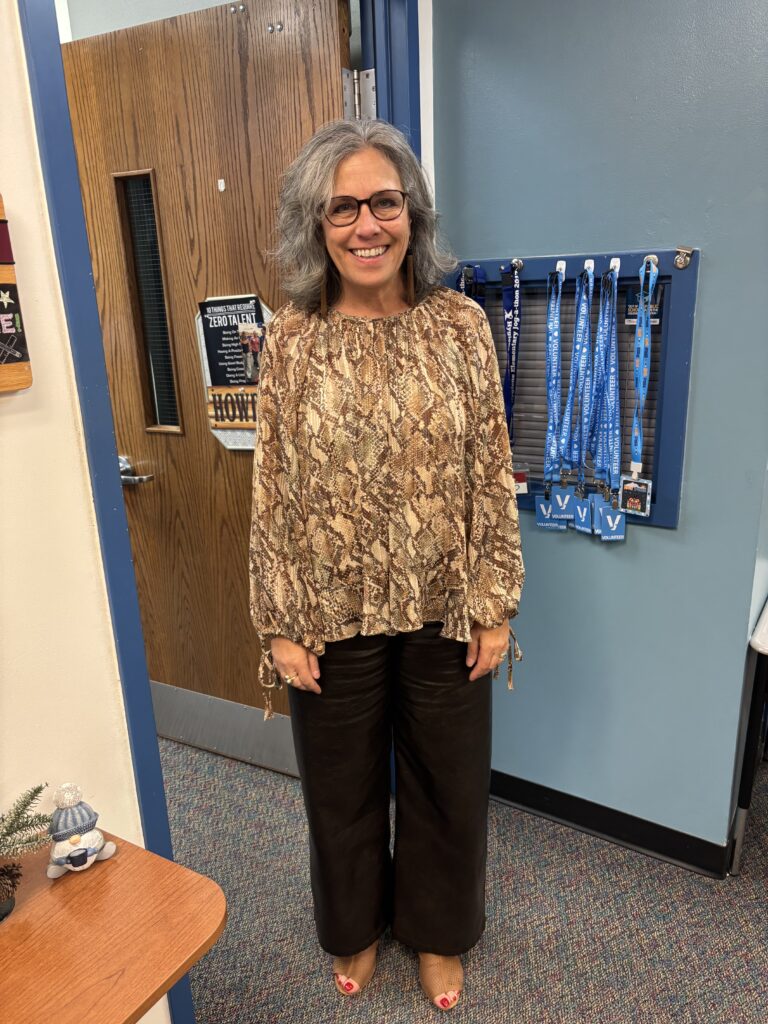 A smiling woman with gray hair wears a patterned blouse and black pants, standing in a colorful office space.