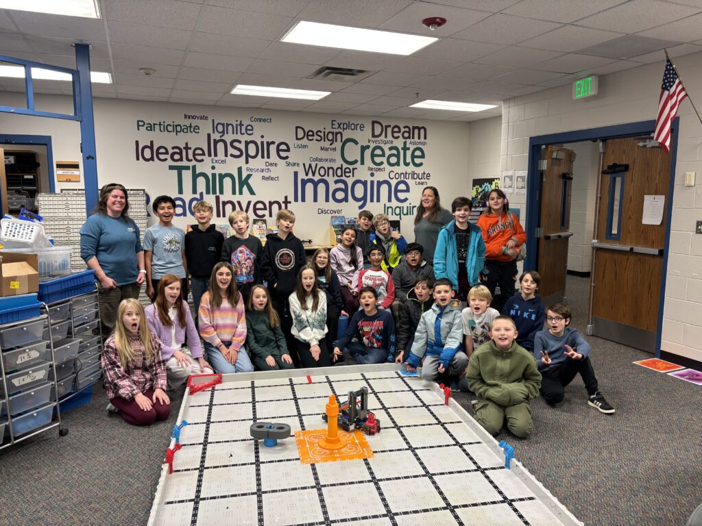 A group of students and a teacher pose together in a classroom with a robotics project on the floor and inspirational words on the wall.