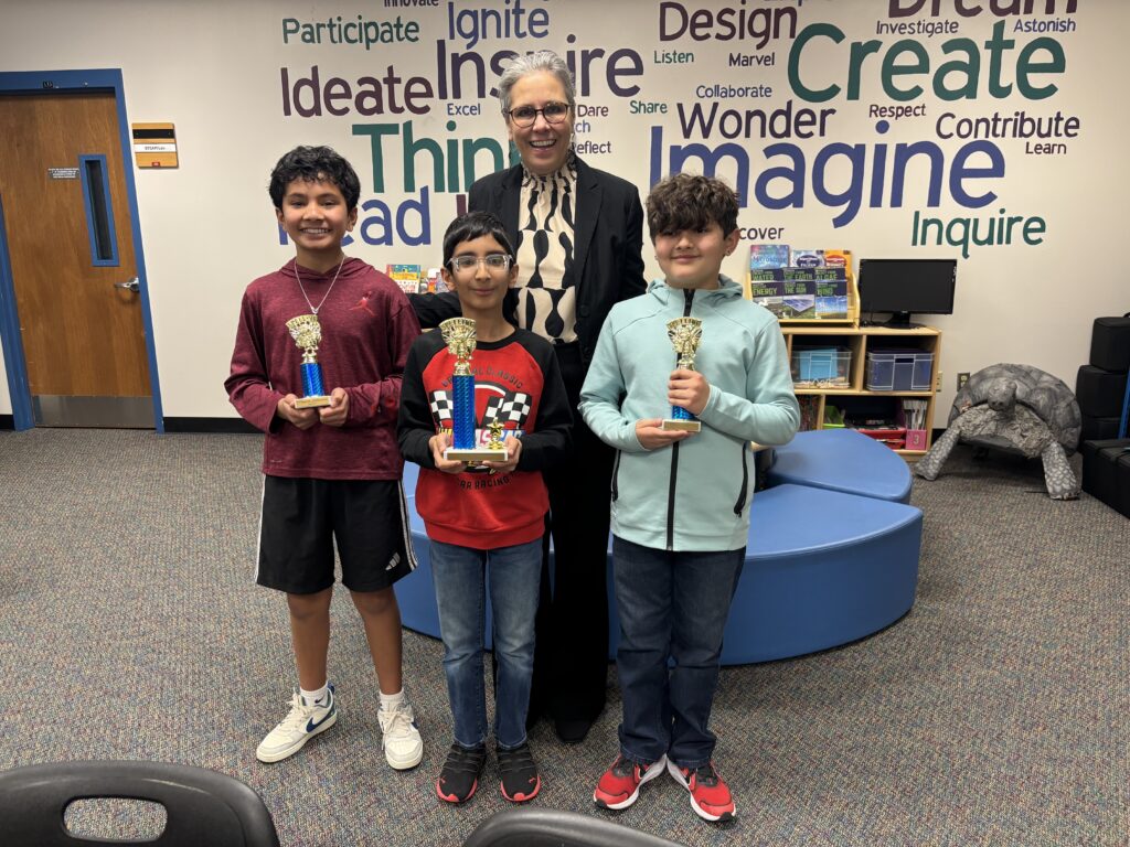 Three children proudly hold trophies while standing with an adult in a colorful classroom with educational words on the wall.
