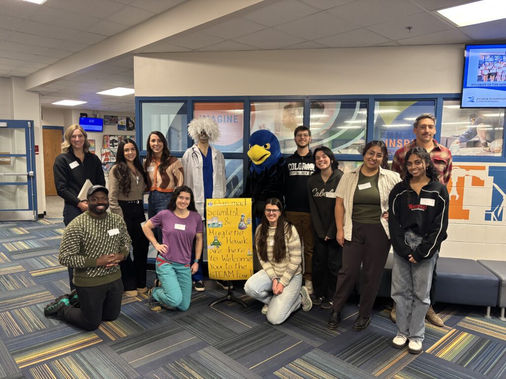 A group of diverse individuals poses with a mascot in a school hallway, welcoming attendees to a STEAM event.