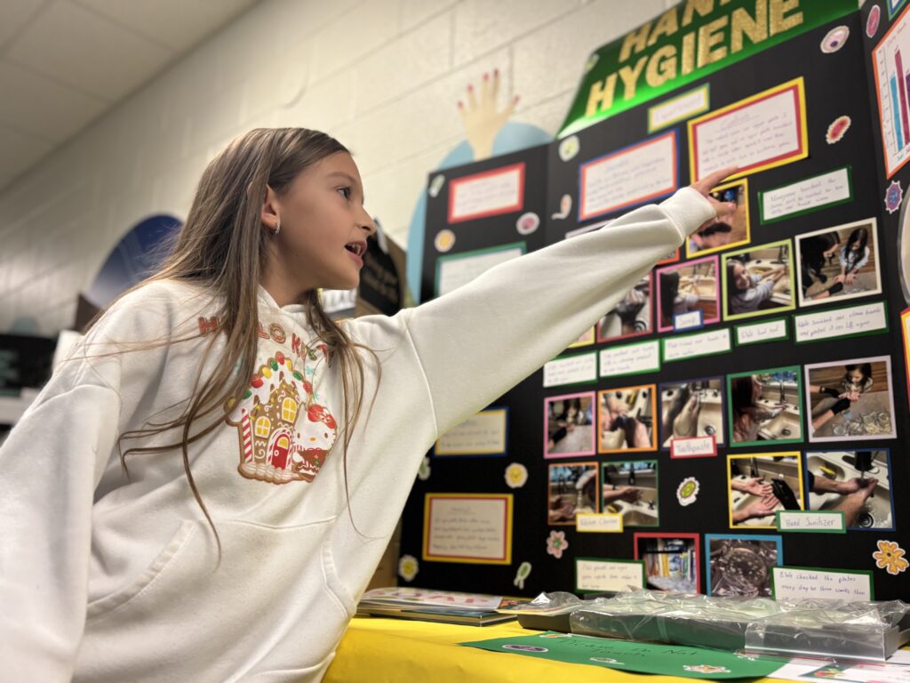 A girl points to a colorful poster about hand hygiene, featuring images and information on proper handwashing techniques.