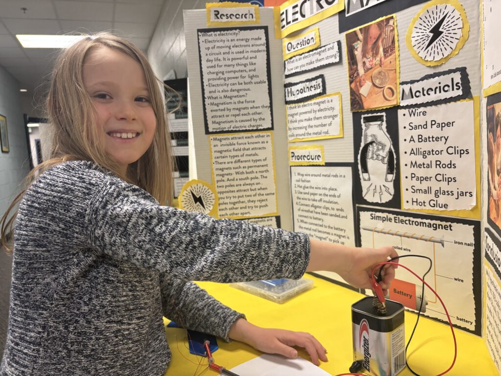 A young girl smiles while demonstrating an electromagnet project, connecting wires to a battery at a science fair display.