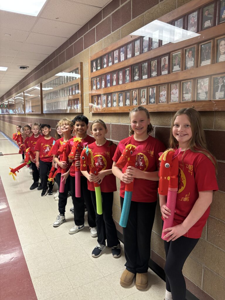 A line of children in red shirts hold colorful, handmade horse puppets in a school hallway.